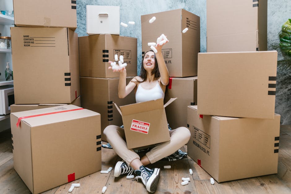 A young woman with long dark hair sitting on a wooden floor surrounded by unpacked cardboard boxes of various sizes, some labeled with 'Fragile'. She is wearing a white sleeveless top, beige pants, an