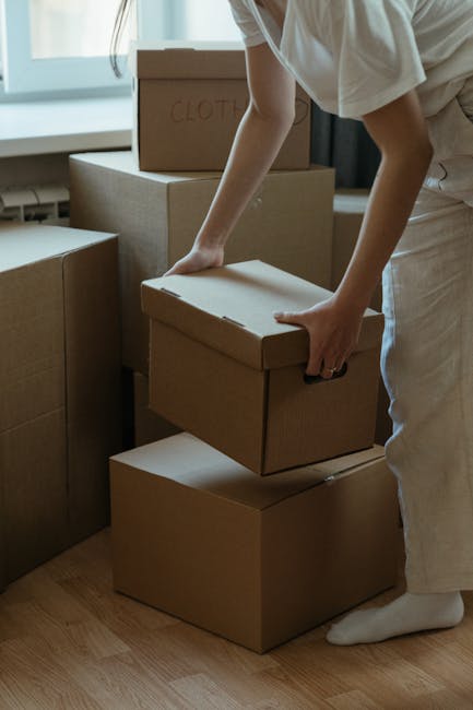 A young woman with long dark hair sitting on a wooden floor surrounded by unpacked cardboard boxes of various sizes, some labeled with 'Fragile'. She is wearing a white sleeveless top, beige pants, an