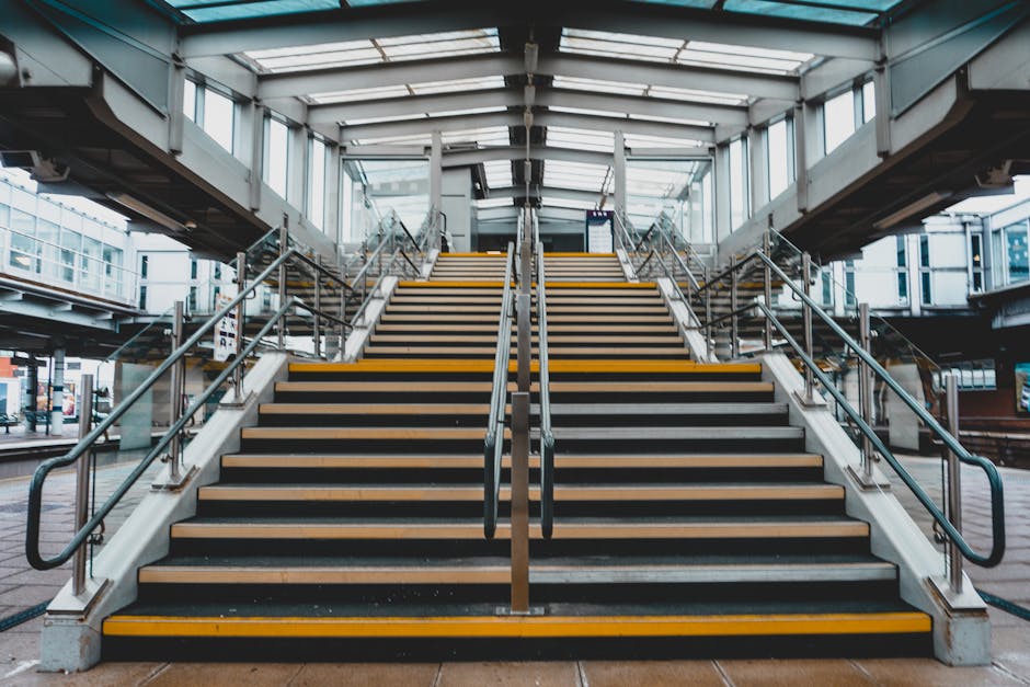 A wide concrete staircase with metal handrails on both sides and yellow tactile strips along the edge of each step, leading up to an enclosed glass and steel pedestrian footbridge. The footbridge has 
