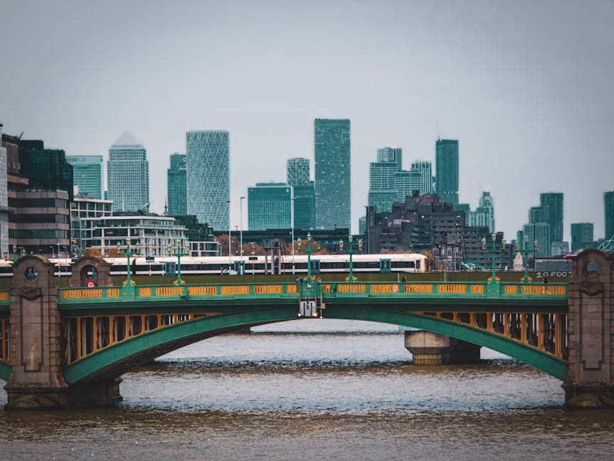 A wide concrete staircase with metal handrails on both sides and yellow tactile strips along the edge of each step, leading up to an enclosed glass and steel pedestrian footbridge. The footbridge has 